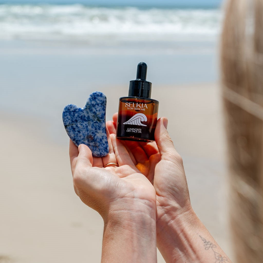 A woman holding the Selkia natural dry face oil serum in sustainable glass jar and the Gua Sha face massage tool with beach ocean and sand blurred background