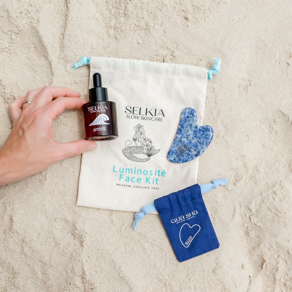 Selkia face kit with a woman hand holding the Selkia natural dry face oil serum accompanied by Gua sha stone face massage tool and blue carrier bag with Selkia kit canvas bag presented on sandy beach background