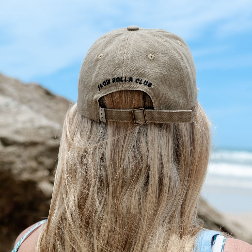 Back of blond woman wearing Selkia naturally dyed tan peak cap with embroidery words - Slow Rolla Club with beach blurred background