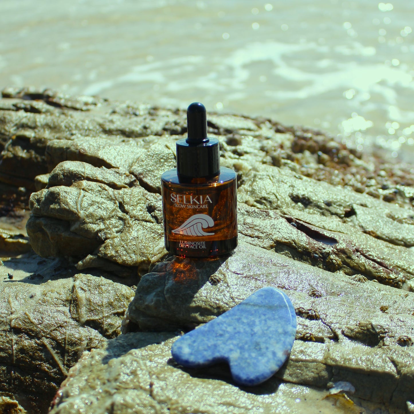 Amber bottle with face oil inside sitting on a rock by the ocean with Gua Sha