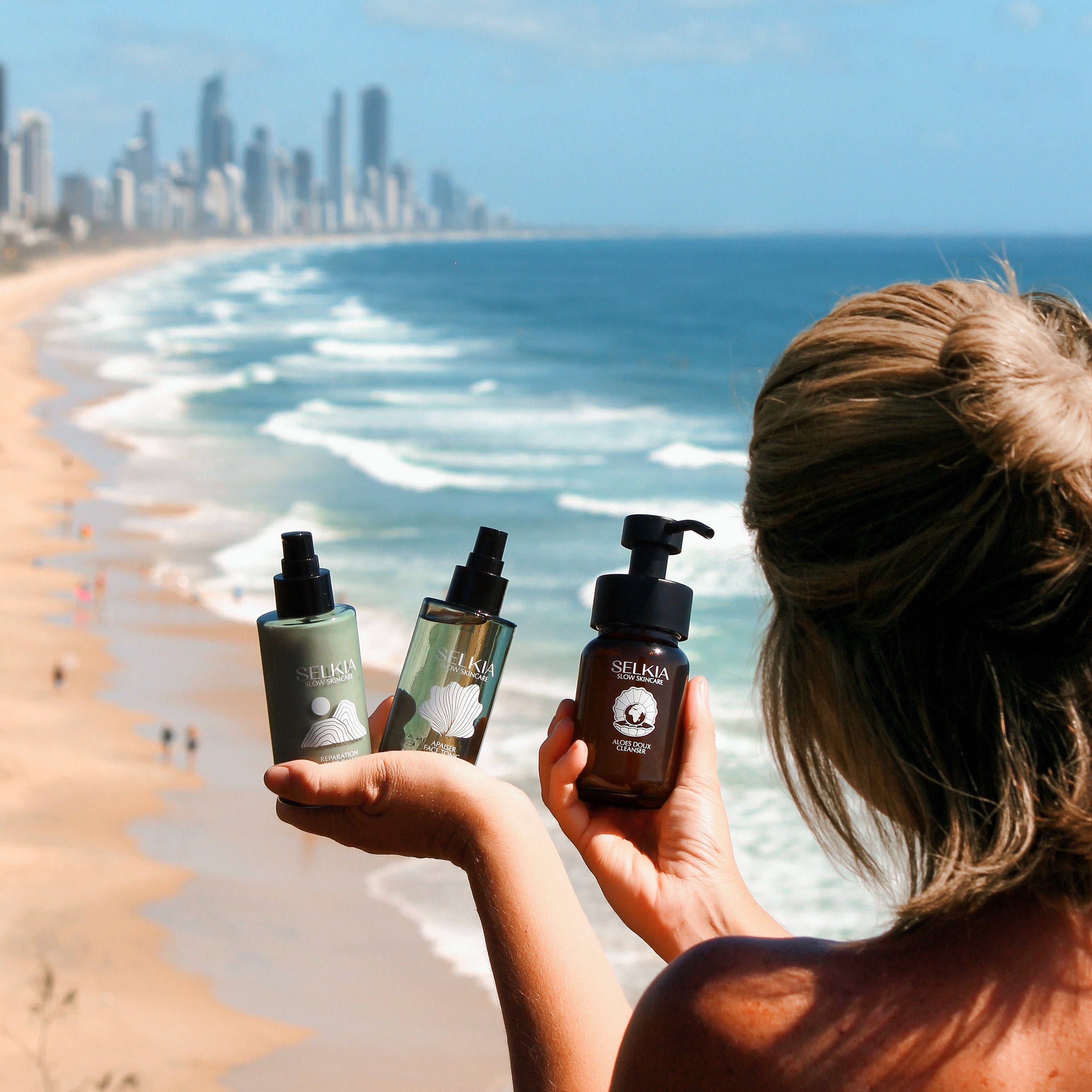 Person holding skincare products on a beach with a city skyline in the background
