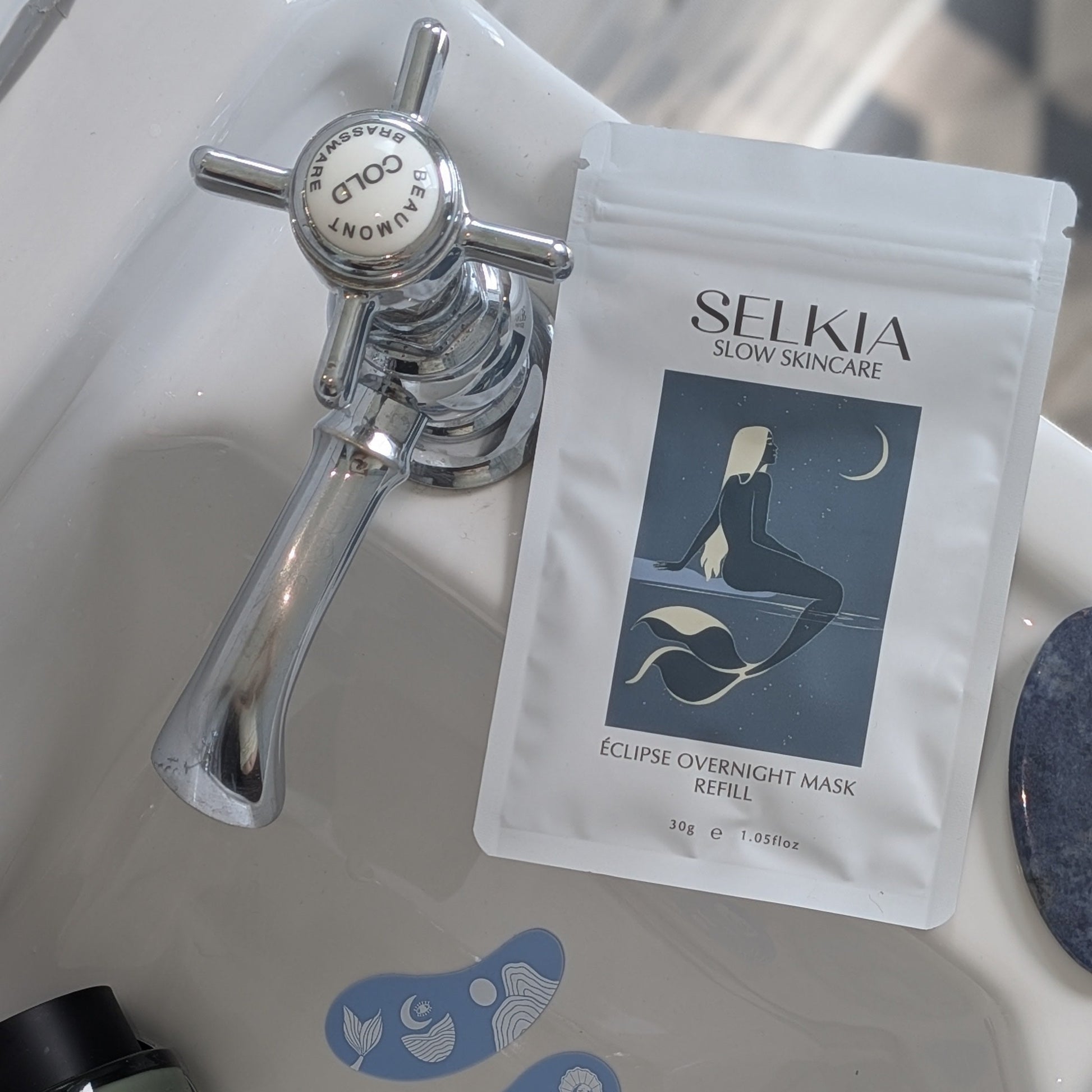 A packet of Selkia Eclipse Overnight Mask placed on a white ceramic sink next to a chrome faucet, with blue mask applicator pads and a small bottle nearby.